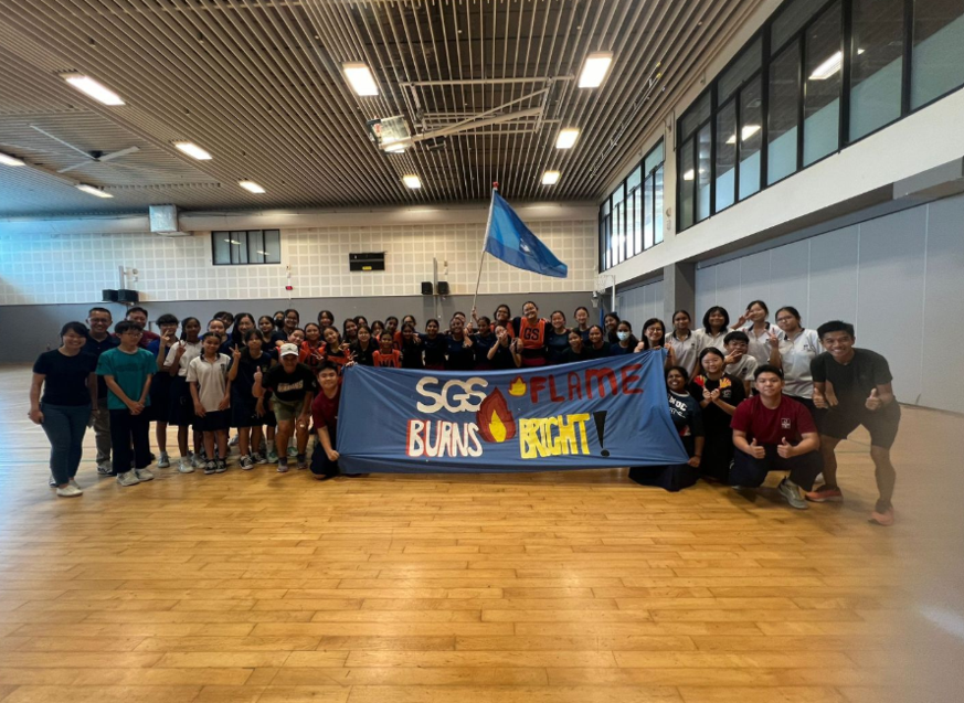 A large group of students and teachers pose together at an indoor court, holding a large blue banner that reads "SGS FLAME BURNS BRIGHT!". A student in the back holds up a blue flag, and the group is celebrating their win at the national school games event.
